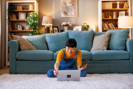 Beautiful Asian woman sitting on the floor using laptop computer and smart phone while working remotely from homeの写真素材