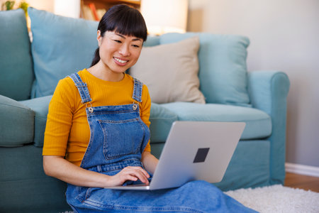 Female freelancer sitting on the floor leaning on the couch holding laptop computer in her lap while working from homeの写真素材