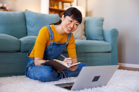 Woman sitting on living room floor, using laptop computer and writing notes in a planner while...
