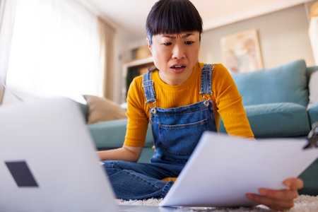 Low angle view of stressed out Japanese woman using laptop computer and reviewing contracts and documentation while working remotely from homeの写真素材
