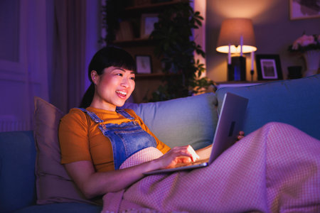 Beautiful mature Asian woman relaxing at home late at night, lying on living room couch havig video call using laptop computer, cheerful smilingの写真素材