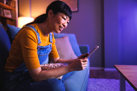 Mature Asian woman sitting at living room couch reading movie review using tablet computer and holding bowl of popcorn in her lap, getting ready for movie night at homeの写真素材