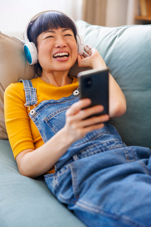 Beautiful Asian woman lying on the couch relaxing and enjoying leisure time at home wearing headset listening to the musicの写真素材