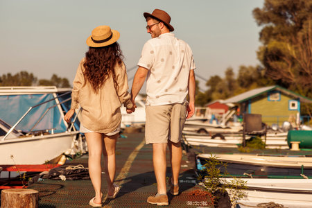 Back view of young couple in love holding hands while taking a walk by the river on the city marina docks, enjoying hot summer day outdoorsの写真素材