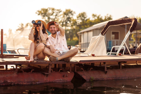 Beautiful young couple in love having fun while on summer vacation, sitting on river docks in city marina, watching birds using binocularsの写真素材