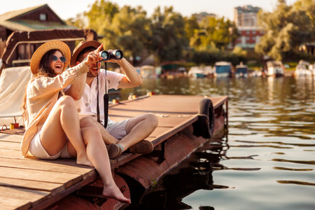 Beautiful young couple in love having fun while on summer vacation, sitting on river docks in city marina, watching birds using binocularsの写真素材