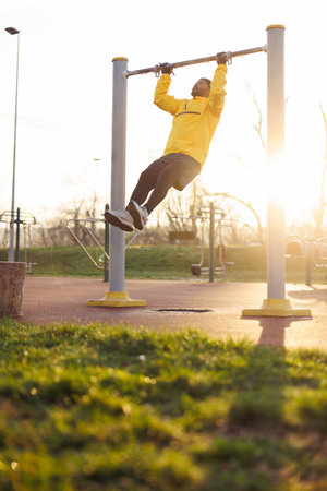 Active athletic young African man doing pull-ups while working out in an outdoor gym in sunrise