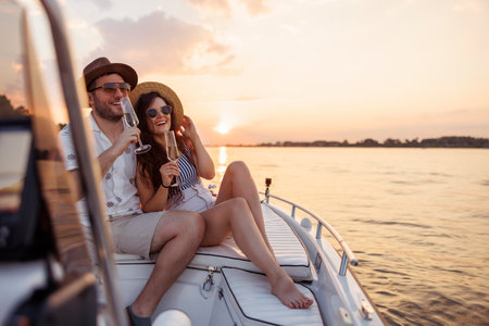 Beautiful young couple in love having fun while on summer vacation, sailing to the sunset on a boat and drinking champagneの写真素材