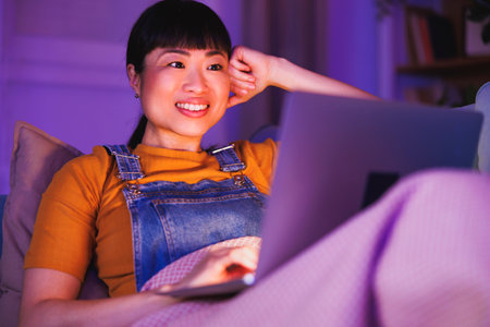 Beautiful mature Asian woman relaxing at home late at night, lying on living room couch using laptop computerの写真素材