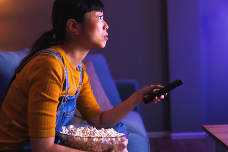 Beautiful Asian woman relaxing at home late at night sitting on living room couch turning on TV using remote control holding a bowl of popcornの写真素材