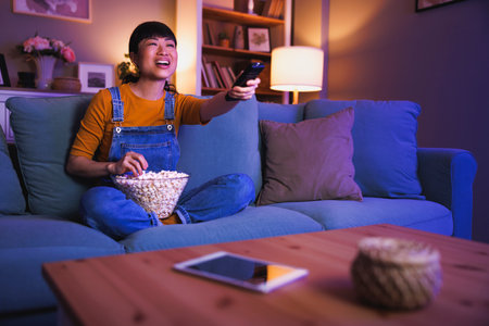 Beautiful Asian woman having fun sitting on living room couch turning on TV using remote control relaxing at home late at nightの写真素材
