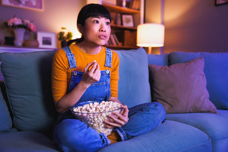 Beautiful Asian woman relaxing at home late at night, eating popcorn and watching documentary film on TV surprisedの写真素材