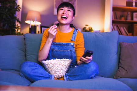 Beautiful Asian woman having fun sitting on living room couch switching channals using remote control and relaxing at home late at night watching TVの写真素材