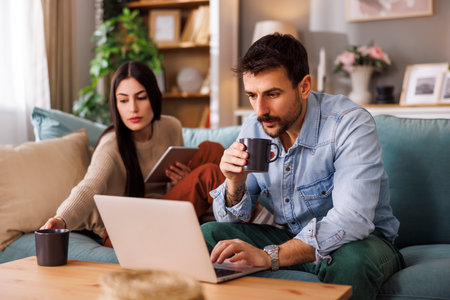 Couple sitting on living room sofa using digital technology devices while working remotely from home, woman using tablet computer while man working on laptop computerの写真素材