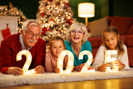 Grandparents celebrating New Years Eve at home with kids, sitting by the Christmas tree, holding illuminative numbers 2025 representing the upcoming New Yearの写真素材