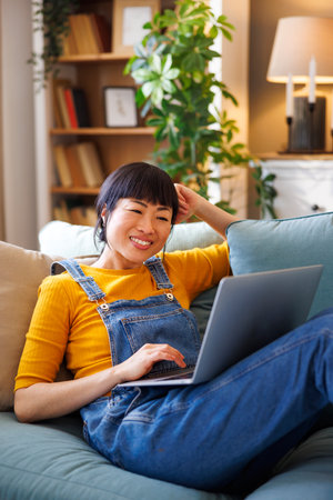 Woman sitting on the couch holding laptop computer in her lap having video call while relaxing at homeの写真素材