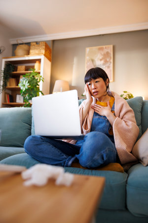 Sick middle aged Japanese woman sitting on living room couch covered with blanket having online doctor's appointment using laptop computerの写真素材