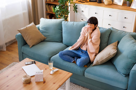 High angle view of middle aged Japanese woman sitting on living room couch covered with blanket while having fever and flu, blowing nose using paper tissueの写真素材