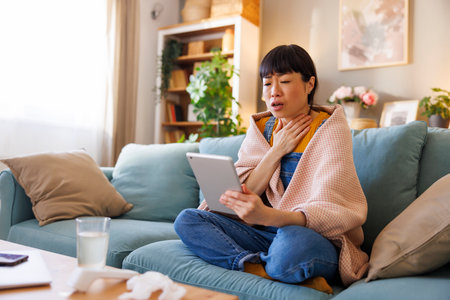 Sick middle aged Japanese woman sitting on living room couch covered with blanket having online doctor's appointment using tablet computerの写真素材