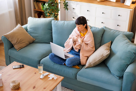 High angle view of sick middle aged Japanese woman sitting on living room couch covered with blanket having online doctor's appointment using laptop computerの写真素材