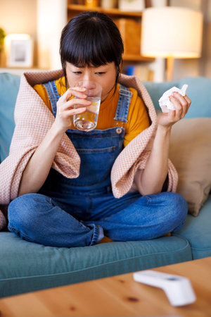 Sick middle aged Japanese woman having fever and flu sitting on living room couch covered with blanket blowing nose and drinking vitamins and waterの写真素材