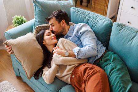 High angle view of beautiful young couple in love lying on living room couch hugging and cuddling while relaxing together at home, man kissing woman on foreheadの写真素材