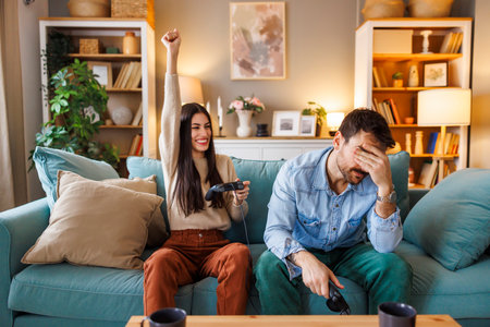 Beautiful young couple in love having fun playing video games while relaxing and enjoying spending leisure time together at home, man unhappy after losingの写真素材