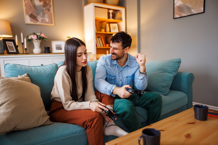 Beautiful young couple in love having fun playing video games while relaxing and enjoying spending leisure time together at home, man celebrating after winningの写真素材
