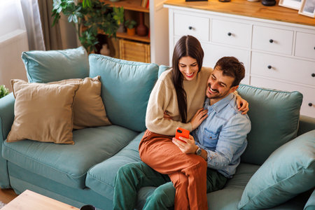 High angle view of beautiful young couple in love hugging and doing online booking for their vacation using smart phone while relaxing at home, woman sitting in man's lapの写真素材