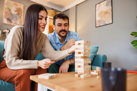 Husband and wife having fun playing block stacking balance game at home, pulling and pushing wooden blocks from sturdy tower in order to keep the balance and not to crash the towerの写真素材