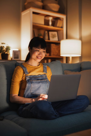 Beautiful woman relaxing at home late at night, sitting on living room couch using laptop computerの写真素材