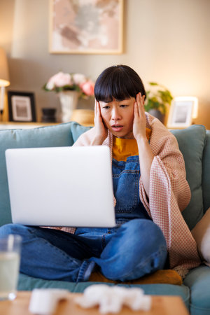 Middle aged Japanese woman having headache sitting on living room couch covered with blanket having online doctor's appointment using laptop computerの写真素材