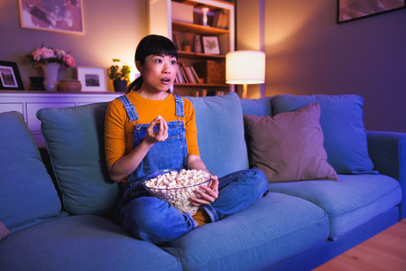 Beautiful Asian woman relaxing at home late at night, eating popcorn and watching TV shockedの写真素材