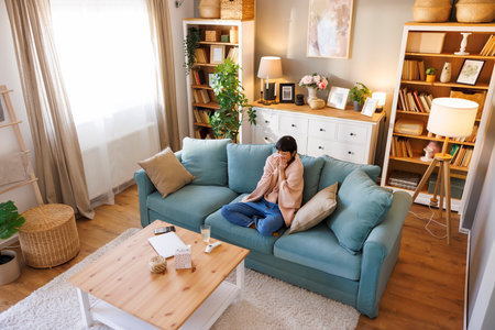 High angle view of middle aged Japanese woman sitting on living room couch covered with blanket while having fever and flu, blowing nose using paper tissueの写真素材