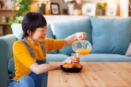 Mature Asian woman pouring a glass of fresh orange juice while having breakfast and relaxing at home in the morningの写真素材