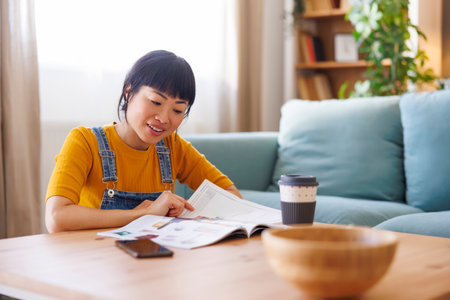 Mature Asian woman relaxing at home reading magazine and drinking coffeeの写真素材