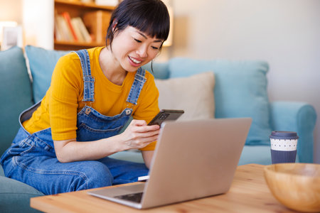 Mature Asian woman working remotely from home using laptop computer, typing text message using smart phone and writing notes in a plannerの写真素材