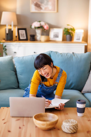Woman sitting on living room floor, working remotely from home using laptop computer and writing notes in a planner while having phone conversationの写真素材