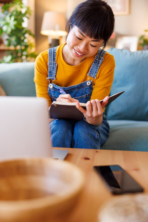 Mature Asian woman having conference call using laptop computer while working remotely from home, taking notes in a plannerの写真素材