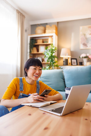 Mature Asian woman working remotely from home using laptop computer, typing text message using smart phone and writing notes in a plannerの写真素材