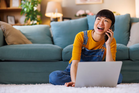 Mature Asian woman having a phone conversation and using laptop computer while working remotely from homeの写真素材