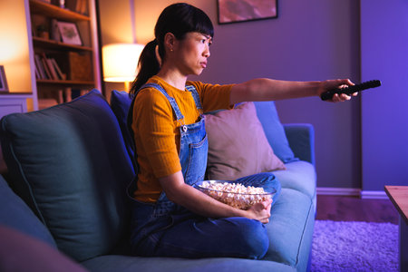 Beautiful Asian woman sitting on living room couch eating popcorn and changing channels using remote control while watching TV at home late at nightの写真素材