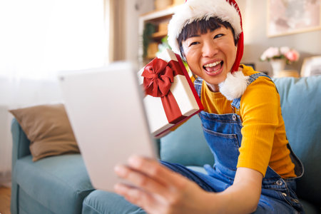 Cheerful Japanese woman wearing Santa hat having video call with friends and family using tablet computer, unboxing Christmas surprise giftの写真素材