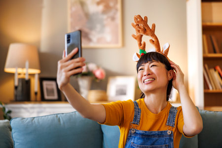 Beautiful cheerful Japanese woman wearing costume reindeer antlers having fun taking selfies using smart phone while celebrating Christmas at homeの写真素材