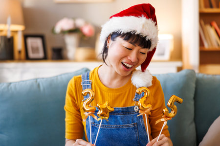 Beautiful cheerful Japanese woman wearing Santa hat having fun holding numbers 2025 representing the...