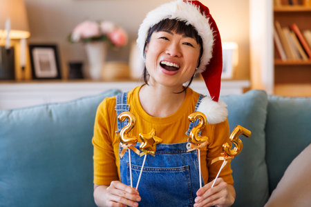 Beautiful cheerful Japanese woman wearing Santa hat having fun holding numbers 2025 representing the upcoming New Year while celebrating Christmas at homeの写真素材