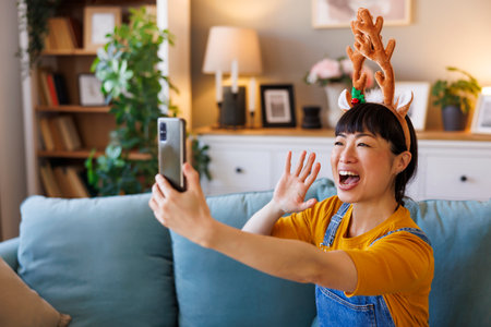 Beautiful cheerful Japanese woman wearing costume reindeer antlers having video call with friends and family using smart phone while celebrating Christmas at homeの写真素材