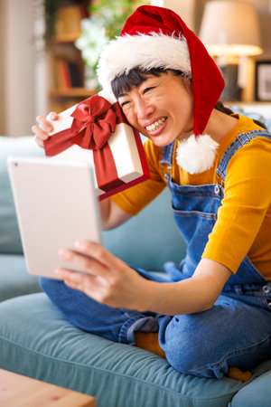 Cheerful Japanese woman wearing Santa hat having video call with friends and family using tablet computer, unboxing Christmas surprise giftの写真素材
