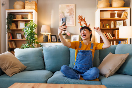 Beautiful cheerful Japanese woman wearing costume reindeer antlers having fun taking selfies using smart phone while celebrating Christmas at homeの写真素材