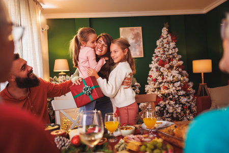 Mother hugging her cute little daughters after getting Christmas surprise present while having Christmas family dinner all together at homeの写真素材